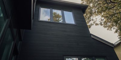 From below of gray sky and green foliage over tall black house with small windows and black wooden walls in daytime