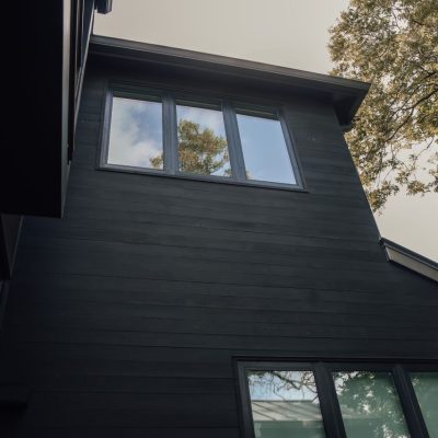 From below of gray sky and green foliage over tall black house with small windows and black wooden walls in daytime